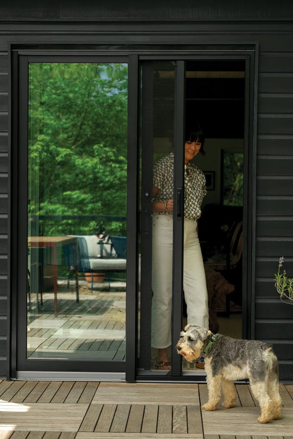 An exterior shot of a woman opening a black 200 Series Narroline® Gliding Patio Door and retractable insect screen to her deck where her dog awaits by a potted plant.